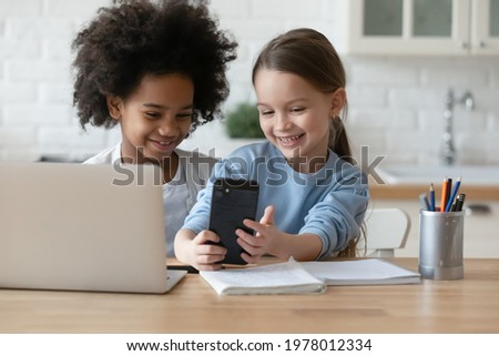 Smiling little multiracial sisters use smartphone talk speak on video call on gadget online. Happy small diverse multiethnic girls children have fun make self-portrait picture on cellphone together.