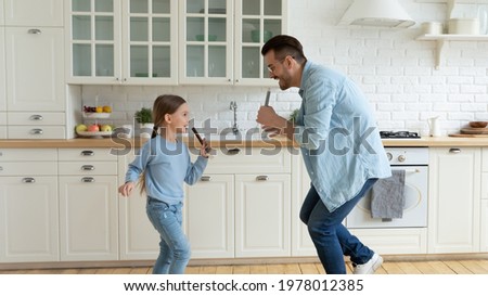 Overjoyed young Caucasian father have fun sing in kitchen appliances with small daughter. Happy dad play and dance at home with little teen girl child on leisure family weekend. Parenthood concept.