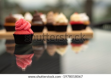 Capcake in the foreground. Background, blurred background with several cupcakes on a board. all of this on a glass table with the images of the sweets reflected.