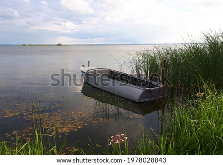 One boat on the water of the lake on a clear summer day. A lone boat on the surface of a lake, tall green grass in the foreground of the photograph, and a blue sky with white clouds.