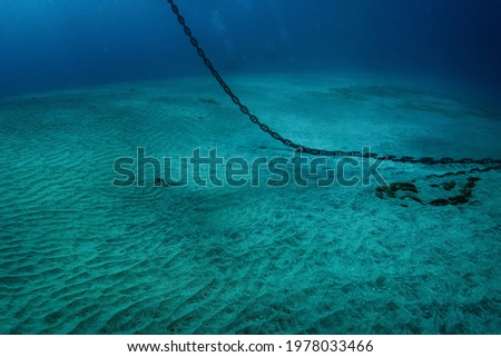 Underwater photo of heavy steel anchor chain that hangs down from water surface and lays on white sand bottom, murky blue water deeper in tropical sea