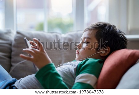 Happy kid wearing earphones listening to music on tablet, Cute boy lying on sofa playing games online on internet in living room, Portrait Child relaxing at home on weekend.