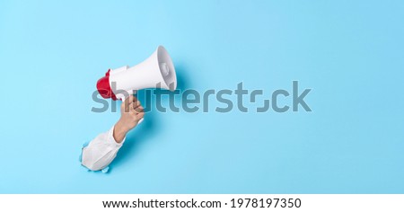 Business Hand holds a megaphone from a hole in the wall on blue background.  hiring, advertising, advertise and Banner concept. Royalty-Free Stock Photo #1978197350