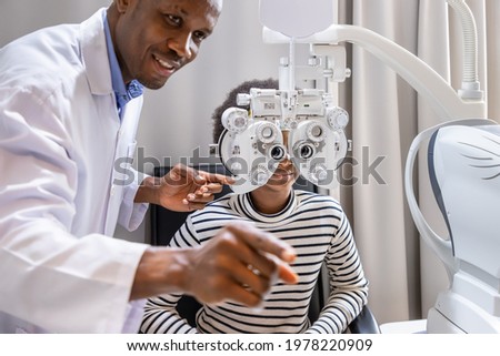 African young woman girl doing eye test checking examination with male man optometrist using phoropter in clinic or optical shop. Eyecare concept. Royalty-Free Stock Photo #1978220909