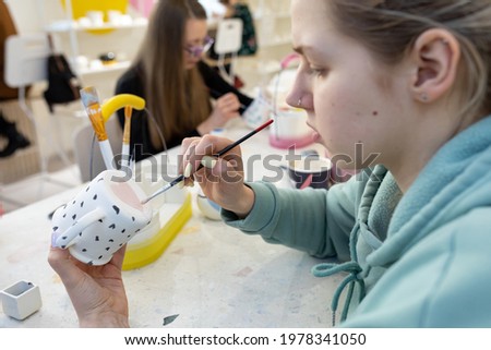 Young woman in casual clothes paint pink heart on back of ceramic cup in ceramic workshop. Learning new skills, improving skills, hobbies. Shallow depth of field, focus on the picture