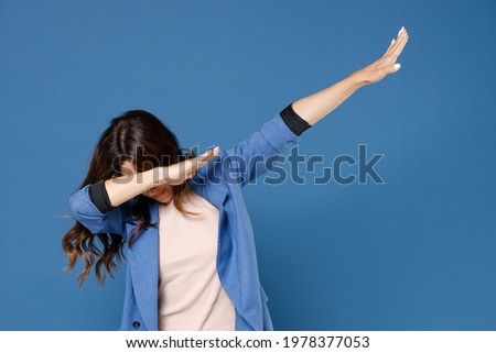 Cheerful funny young brunette woman 20s in basic jacket standing doing dab hip hop dance hands gesture, youth sign hiding and covering face isolated on bright blue colour background, studio portrait