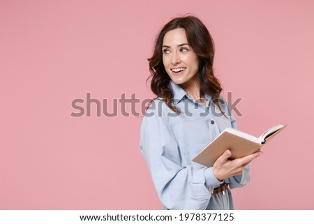 Side view of smiling attractive young brunette woman 20s wearing casual blue shirt dress posing hold in hands reading book looking aside isolated on pastel pink colour background, studio portrait