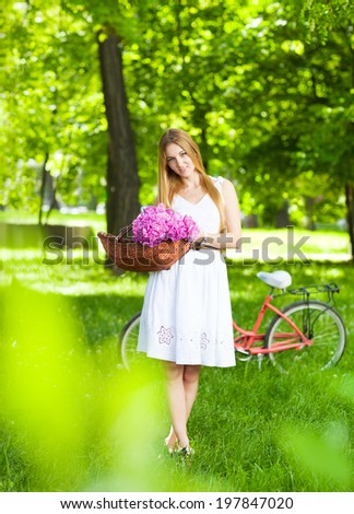 Beautiful blond woman wearing a nice dress having fun in park with bicycle carrying a beautiful basket full of peony flowers. Vintage scenery