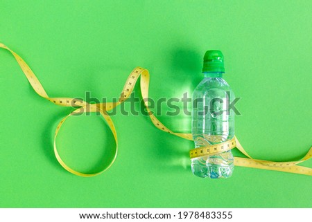 A bottle of still water and a measuring tape on a green background. The concept of losing weight, healthy lifestyle, fasting. Drinking regime during exercise, sports. Duncan's diet. World Obesity Day. Royalty-Free Stock Photo #1978483355