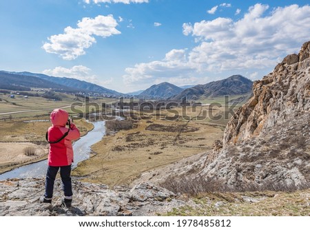 a little girl, a child in an autumn warm jacket stands and takes pictures with a camera of a landscape with a river, standing on a stone of a high mountain