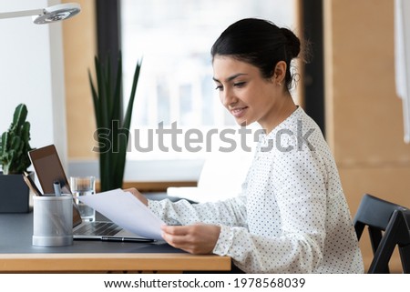 Smiling confident Indian businesswoman reading document, working on laptop, sitting at desk, young woman employee or student typing, holding paper, preparing financial report or research project