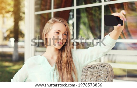 Close up of young blonde woman taking a selfie picture by smartphone sitting at a table in a cafe