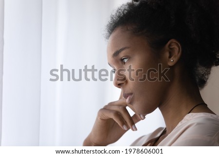 Close up head shot side view young african american thoughtful woman standing near window, looking in distance, feeling puzzled with personal problems, pondering about difficulties, copy space. Royalty-Free Stock Photo #1978606001