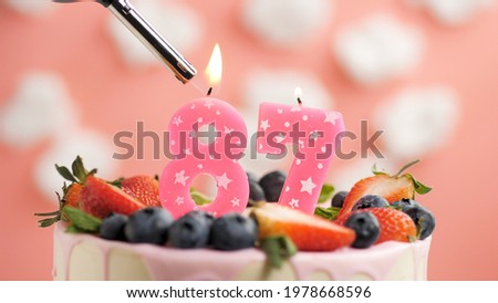 Birthday cake number 87, pink candle on beautiful cake with berries and lighter with fire against background of white clouds and pink sky. Close-up