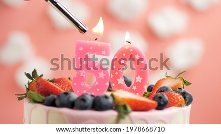 Birthday cake number 50, pink candle on beautiful cake with berries and lighter with fire against background of white clouds and pink sky. Close-up