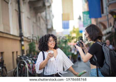 Tourist in the city.  Smiling woman standing on the street with cup of coffee. Woman taking photo of her friend. Focus is on background. 