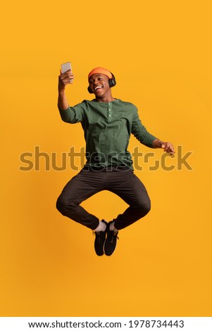 Joyful Black African Guy With Wireless Headphones And Smartphone Jumping In Air, Positive Black Hipster Man Listening Music And Having Fun Over Yellow Studio Background, Full-Length, Copy Space