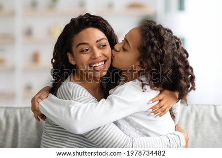 Loving african american school girl with curly hair kissing her beautiful mother, closeup portrait, copy space. Adorable black family young woman and her daughter bonding at home