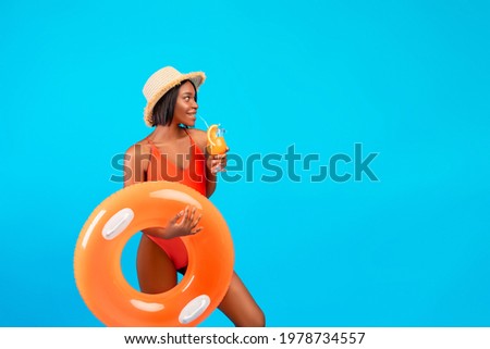 Summertime chill. Attractive black woman in swimwear holding inflatable ring and drinking fruit cocktail on blue studio background, copy space. Cool young lady having fun on tropical vacation