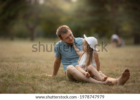 Dad and daughter in summer park sit on the grass. Girl hugs and kisses the dad, father smiles and happy. Family leisure concept, nature walk, family care and values, fatherhood, fathers day