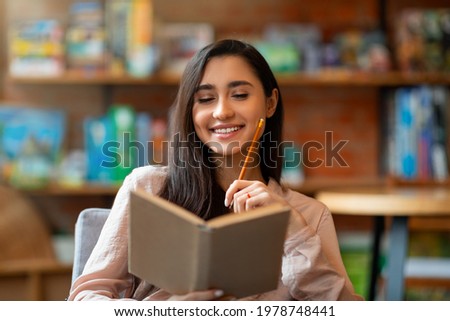 Happy laitin woman thinking about future plans, writing list to do in notepad or planning her day while sitting in cafeteria. Excited arab lady taking notes at notebook