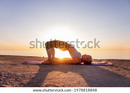 Young blonde woman in sportswear performs yoga asanas, exercises on the seashore at sunrise. The girl in sports, works out breathing, meditates, performs Half Bridge Pose. Healthy mind in fit body Royalty-Free Stock Photo #1978818848
