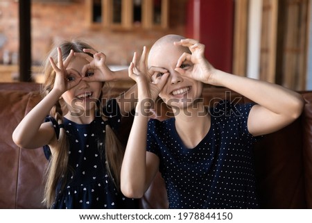 Happy mom and little daughter relaxing, playing and having fun at home. making faces with finger glasses, smiling and laughing. Optimistic mother enjoying leisure time with kid despite cancer
