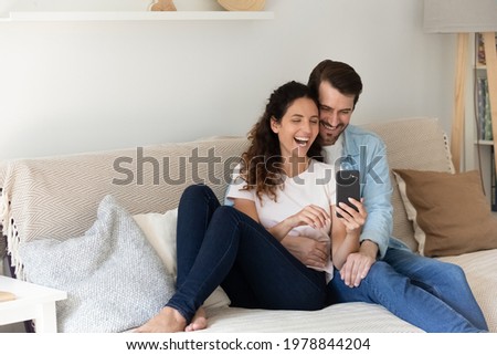 Happy bonding young family couple resting on couch, watching funny photos in social network, having fun entertaining playing mobile games together. Laughing spouses holding web camera video call.