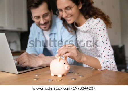 Happy millennial family couple putting coins in piggybank, planning vacation or investments together, saving money for life insurance, managing future expenditures together using computer apps. Royalty-Free Stock Photo #1978844237