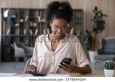 Happy young african ethnic woman in glasses calculating domestic utility bills taxes, managing monthly budget, paying for services online in mobile shopping application, satisfied with enough money. Royalty-Free Stock Photo #1978844264
