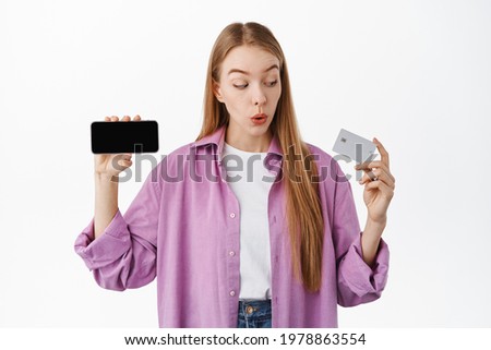 Young woman bank client, looking amazed at credit card and showing mobile phone application, smartphone screen flipped horizontal, standing over white background