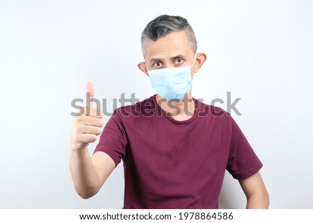 Portrait of an Asian man in casual tshirt, wearing medical mask, showing an okay sign, agreeing with you, praising a good choice, standing on a white background.