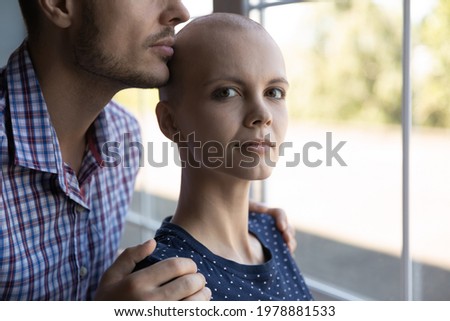 Portrait of serious determined young woman fighting against cancer with love and support of husband or boyfriend. Man embracing ill wife, standing at window, looking at camera. Oncology, hope concept
