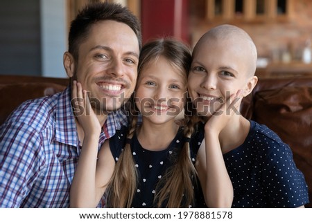 Family portrait of happy little girl with smiling dad and optimistic hairless ill mom. Head shot of young couple and child sitting on couch at home, looking at camera, smiling. Oncology concept