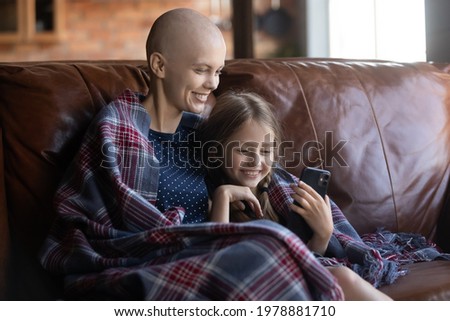 Happy ill mom and little daughter wrapped in plaid relaxing on coach, using mobile phone for playing online game or video call and virtual talk to dad. Mother with cancer spending time with kid