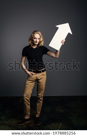 A handsome young guy with long blonde hair smiles and holds on a white arrow directed to the right on a gray background.