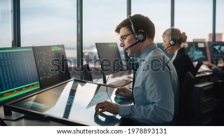 Male Air Traffic Controller with Headset Talk on a Call in Airport Tower. Office Room is Full of Desktop Computer Displays with Navigation Screens, Airplane Departure and Arrival Data for the Team. Royalty-Free Stock Photo #1978893131