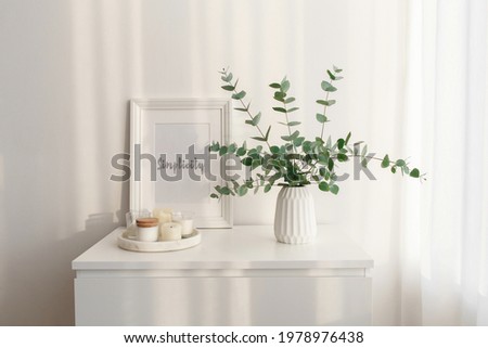 Frame with text SIMPLICITY, bouquet of eucalyptus branches in vase and tray with candles standing on the chest of drawers in the morning sun beams. Minimalistic home decor. White stylish interior.