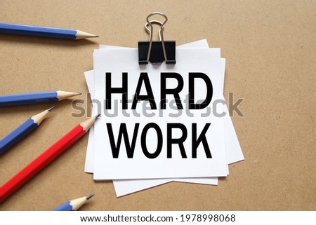 HARD WORK. text on the sticker. paper is clamped with a paper clip. paper on a wooden background near different pencils