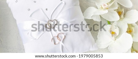 banner with white pillow for wedding rings with rings and rhinestone hearts near white orchid on a white textured background. Wedding or engagement concept. soft focus