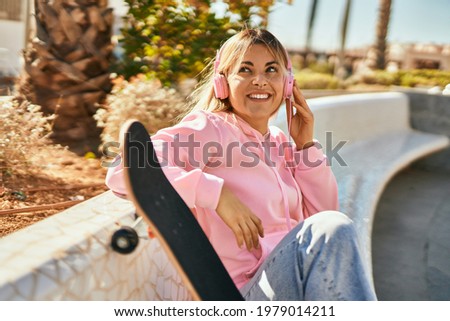 Young blonde skater girl smiling happy using headphones sitting at the park.