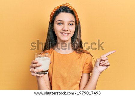 Young brunette girl drinking a glass of milk smiling happy pointing with hand and finger to the side 