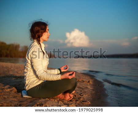 Alone girl on the sandy beach near the river does yoga and meditation on a background of blue sky and white clouds