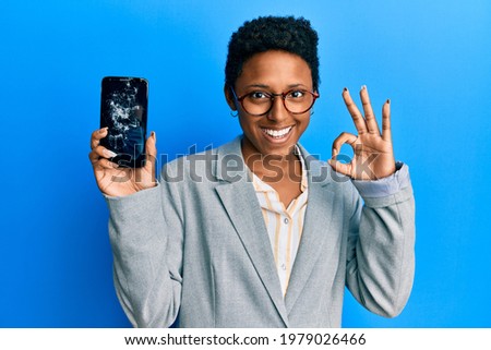 Young african american girl holding broken smartphone showing cracked screen doing ok sign with fingers, smiling friendly gesturing excellent symbol 