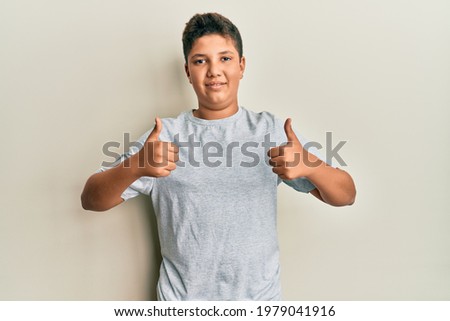 Teenager hispanic boy wearing casual grey t shirt success sign doing positive gesture with hand, thumbs up smiling and happy. cheerful expression and winner gesture. 