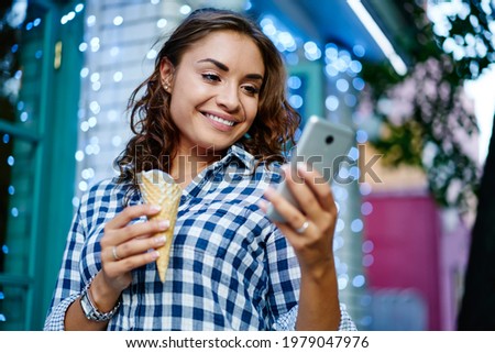 Cheerful Caucaisan woman with ice cream reading text publication in social networks using smartphone application during travel trip, happy millennial browsing social media and mobile website