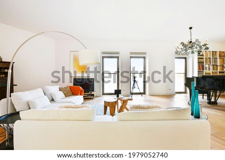 White sofas and tables on carpet against piano under chandelier in spacious living room decorated with art paintings