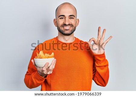 Young bald man holding nachos potato chips doing ok sign with fingers, smiling friendly gesturing excellent symbol 