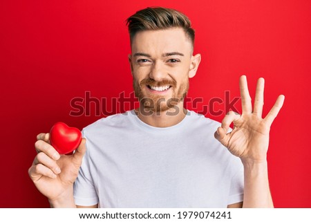 Young redhead man holding heart doing ok sign with fingers, smiling friendly gesturing excellent symbol 