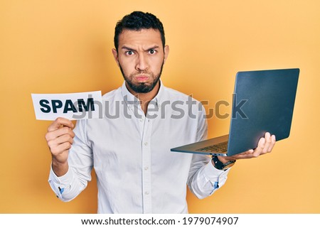 Hispanic man with beard working using computer laptop holding spam banner puffing cheeks with funny face. mouth inflated with air, catching air. 